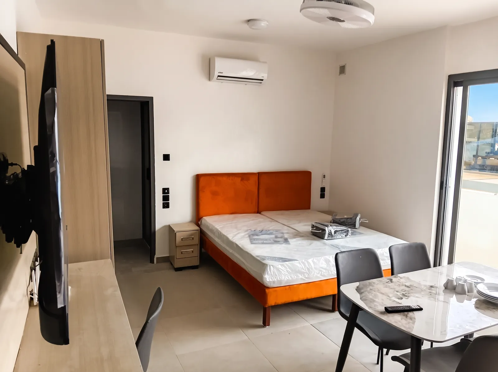 Chambre avec lit à tête de lit orange, climatiseur mural, télévision et table, ouverte sur l'extérieur.